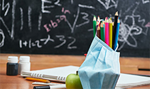 Photo of a teacher's desk with colored pencils, apple, notebook, pencil, and surgical mask with blackboard in background.
