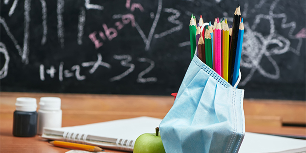 Photo of a teacher's desk with colored pencils, apple, notebook, pencil, and surgical mask with blackboard in background.